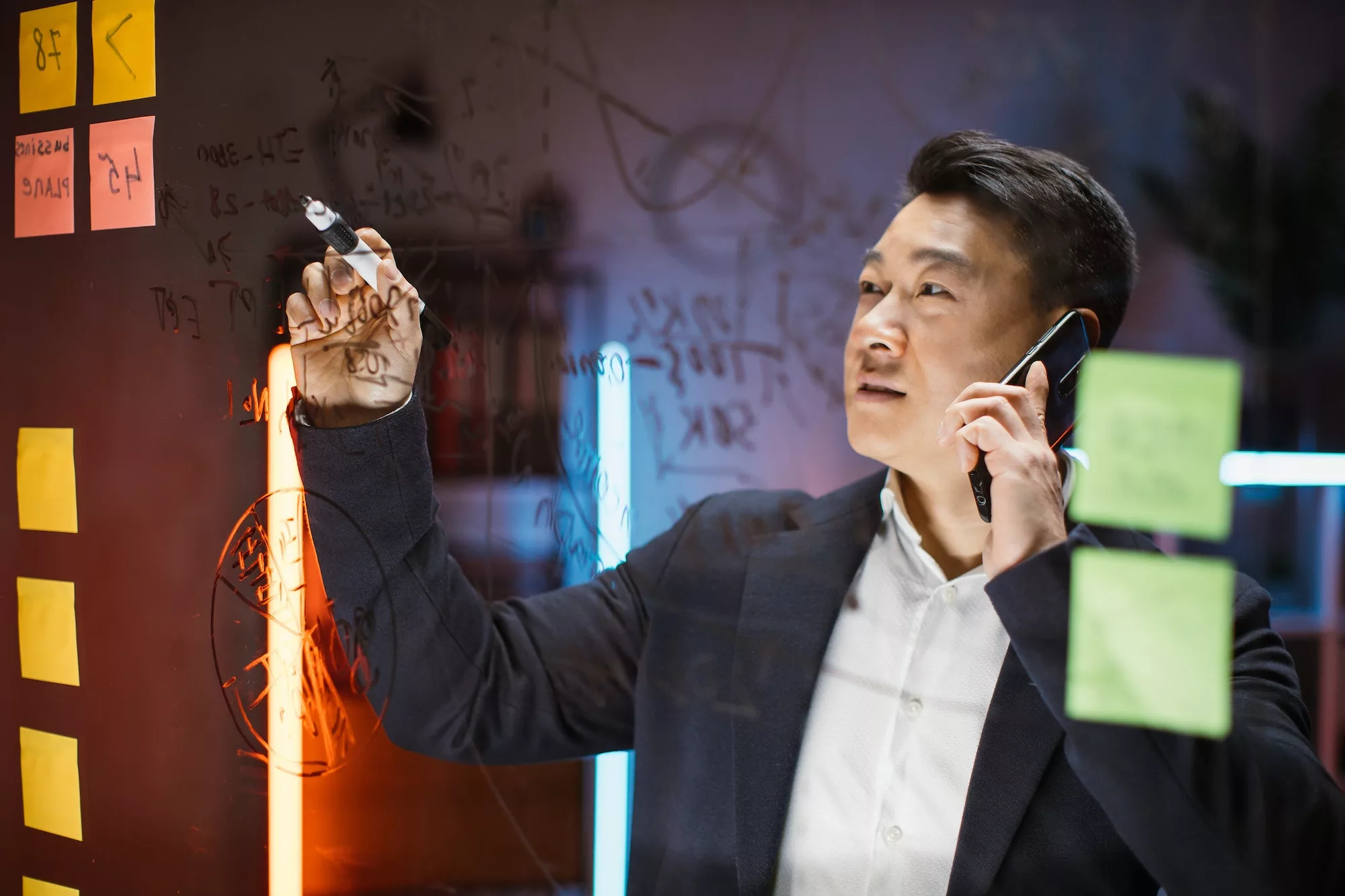 Focused Asian male ceo writing on glass board with sticky notes and talking phone